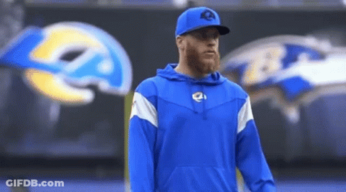A Man Wearing A Blue Hoodie And A Baseball Cap Stands In Front Of A Large Rams Logo . GIF