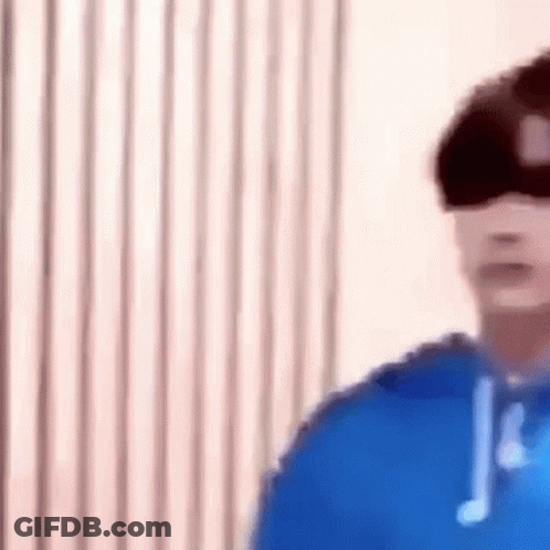 A Man Wearing A Blue Hoodie And Headphones Is Standing In Front Of A Window . GIF