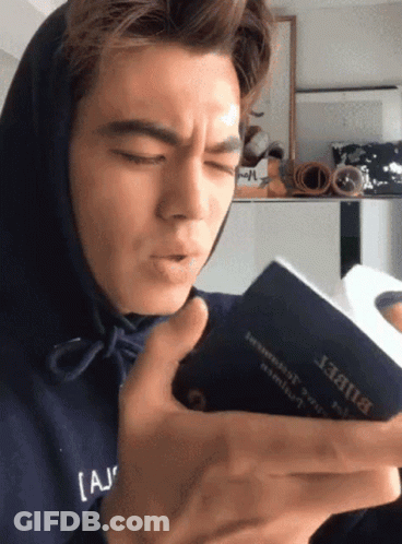 A Man Wearing A Blue Hoodie Is Reading A Book With The Word Bible On It GIF