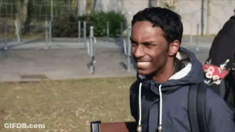 A Man Wearing A Blue Jacket And A Backpack Is Smiling While Standing In A Park . GIF