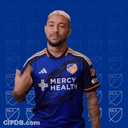 A Man Wearing A Blue Mercy Health Jersey Stands In Front Of A Blue Background With Mls Logos GIF