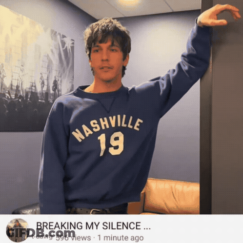 A Man Wearing A Blue Nashville 19 Sweatshirt Leaning Against A Wall GIF