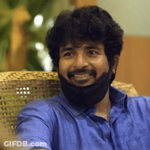 A Man Wearing A Blue Shirt And A Black Face Mask Smiles While Sitting In A Chair GIF