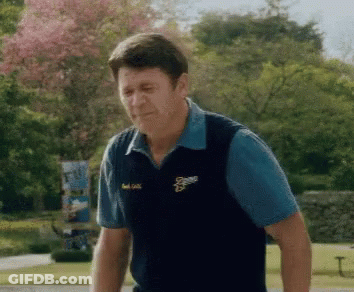 A Man Wearing A Blue Shirt And A Black Vest With The Letter S On It Is Standing In A Park . GIF