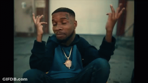 A Man Wearing A Blue Shirt And A Gold Necklace Is Sitting On The Ground With His Hands Up GIF