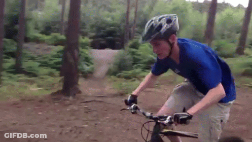 A Man Wearing A Blue Shirt And A Helmet Is Riding A Bike On A Dirt Road GIF