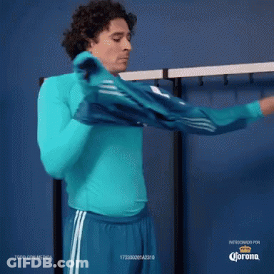 A Man Wearing A Blue Shirt And Blue Shorts Is Standing In A Locker Room With Corona Written On The Bottom GIF