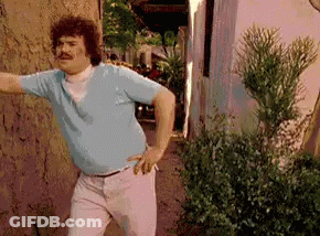A Man Wearing A Blue Shirt And White Pants Is Standing In Front Of A Wall . GIF