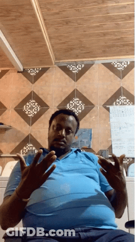 A Man Wearing A Blue Shirt Is Sitting In Front Of A Wall With A Calendar On It GIF