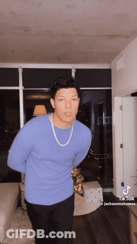 A Man Wearing A Blue Sweater And A Gold Chain Is Standing In A Living Room .. GIF