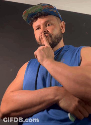 A Man Wearing A Blue Tank Top And A Hat Holds His Finger To His Nose GIF