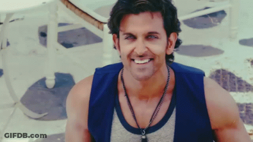 A Man Wearing A Blue Vest And A Necklace Smiles GIF