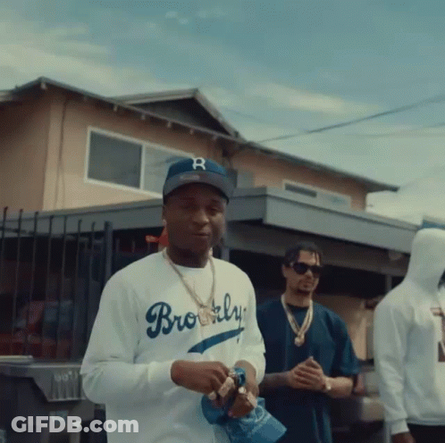 A Man Wearing A Brooklyn Baseball Cap Stands In Front Of A House GIF
