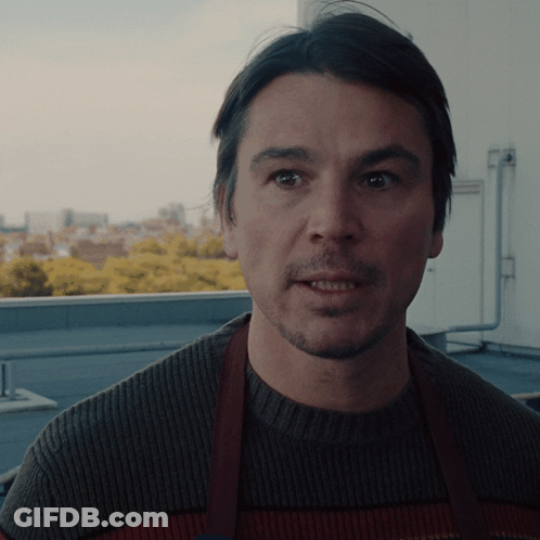 A Man Wearing A Brown Apron And A Sweater Looks At The Camera GIF