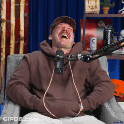 A Man Wearing A Brown Hoodie Is Laughing In Front Of A Microphone With The Letter C On It GIF
