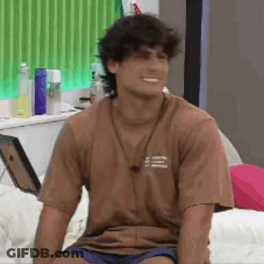 A Man Wearing A Brown Shirt Is Sitting On A Bed . GIF