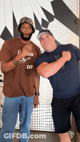 A Man Wearing A Brown Shirt That Says ' Foods By Us ' On It Stands Next To Another Man GIF