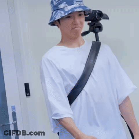 A Man Wearing A Bucket Hat And A White Shirt Is Standing Next To A Tripod . GIF