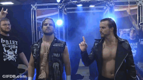A Man Wearing A Bullet Club Shirt Stands Next To Another Man GIF