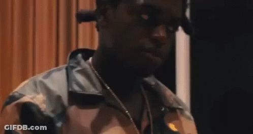 A Man Wearing A Camouflage Jacket And A Necklace Is Standing In Front Of A Curtain . GIF