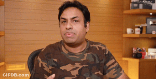 A Man Wearing A Camouflage Shirt Is Sitting In Front Of A Shelf With Geekkiranjit Written On It GIF