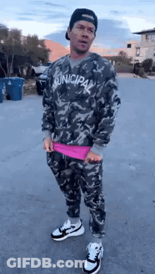 A Man Wearing A Camouflage Sweater And Pants Is Standing On A Street . GIF