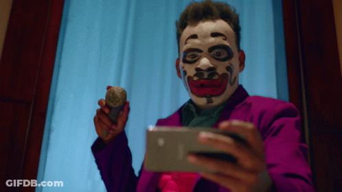 A Man Wearing A Clown Mask Is Holding A Rock And Looking At A Cell Phone GIF