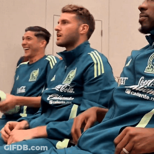 A Man Wearing A Coca Cola Shirt Sits Next To Another Man Wearing A Caliente Shirt GIF