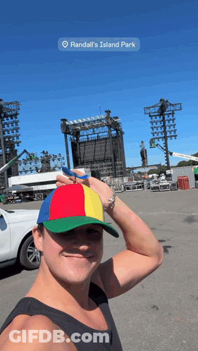 A Man Wearing A Colorful Hat Is Standing In Front Of A Large Stage At Randall 'S Island Park GIF