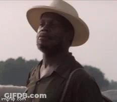 A Man Wearing A Cowboy Hat And A Backpack Is Standing In Front Of A Body Of Water . GIF