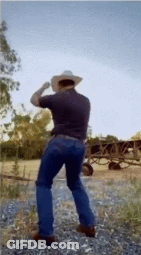 A Man Wearing A Cowboy Hat And Jeans Is Standing In A Field . GIF