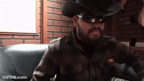 A Man Wearing A Cowboy Hat And Sunglasses Is Sitting In A Chair GIF