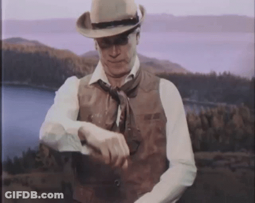 A Man Wearing A Cowboy Hat And Vest Looks At His Watch GIF