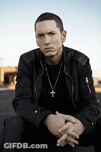 A Man Wearing A Cross Necklace Is Sitting With His Hands Folded GIF