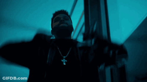 A Man Wearing A Cross Necklace Is Standing In A Dark Room GIF