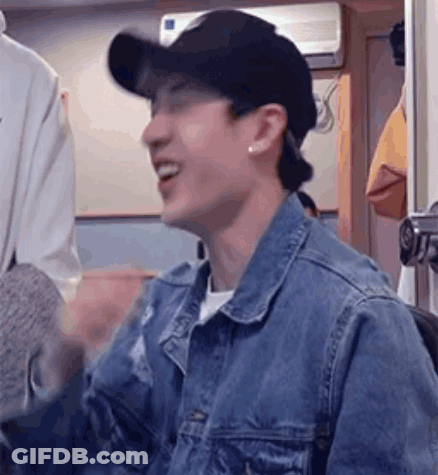A Man Wearing A Denim Jacket And A Baseball Cap Is Smiling And Laughing . GIF