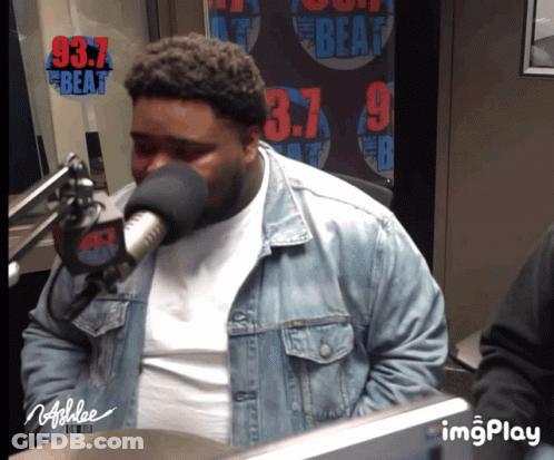 A Man Wearing A Denim Jacket Is Speaking Into A Microphone In Front Of A Sign That Says 97.7 The Beat GIF