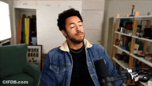 A Man Wearing A Denim Jacket Stands In Front Of A Microphone With A Work Hard Poster On The Wall Behind Him GIF