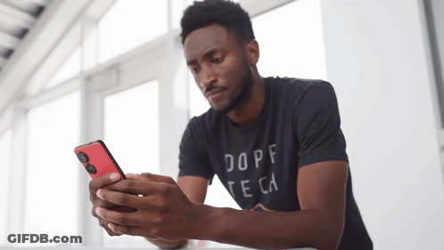 A Man Wearing A Dope Te Ca Shirt Looks At His Cell Phone GIF