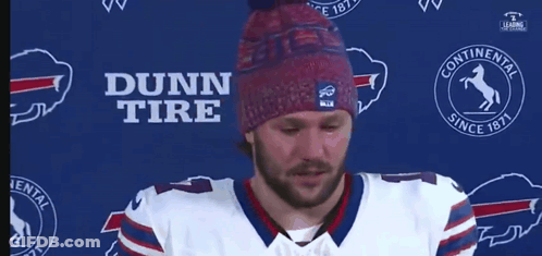 A Man Wearing A Dunn Tire Hat Stands In Front Of A Blue Background GIF