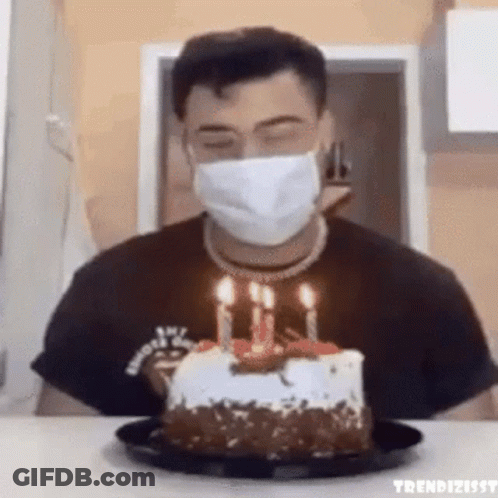 A Man Wearing A Face Mask Is Blowing Out Candles On A Cake . GIF