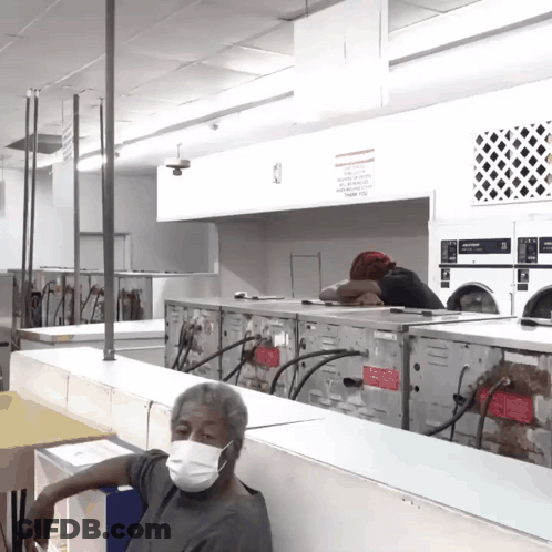 A Man Wearing A Face Mask Sits In A Laundromat Surrounded By Washers And Dryers GIF