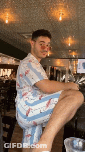 A Man Wearing A Flamingo Shirt And Shorts Sits On A Bar Stool GIF