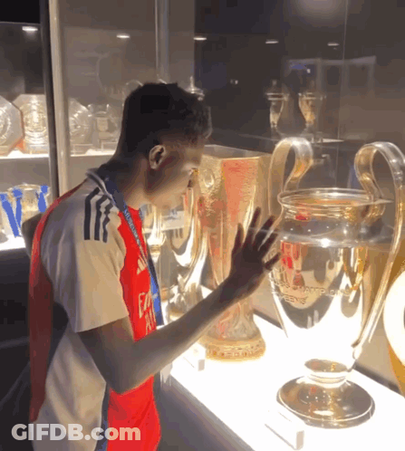 A Man Wearing A Fly Emirates Shirt Touches A Trophy In A Glass Case GIF