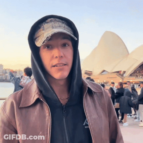 A Man Wearing A George Johnston Hat Stands In Front Of A Building GIF
