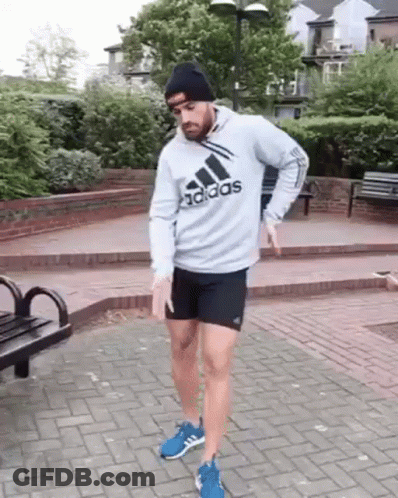A Man Wearing A Gray Adidas Hoodie And Black Shorts Is Standing On A Brick Sidewalk . GIF