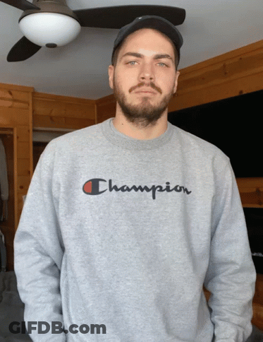 A Man Wearing A Gray Champion Sweatshirt Stands In Front Of A Ceiling Fan GIF