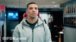 A Man Wearing A Gray Nike Hoodie Is Standing In A Hallway . GIF