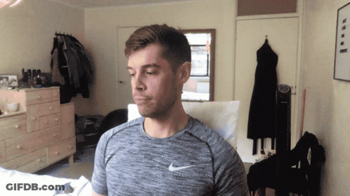 A Man Wearing A Gray Nike Shirt Stands In A Bedroom GIF