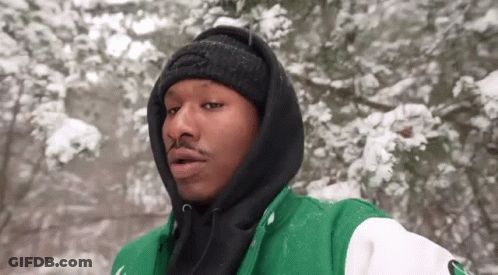 A Man Wearing A Green And Black Jacket And A Black Beanie Is Standing In The Snow . GIF